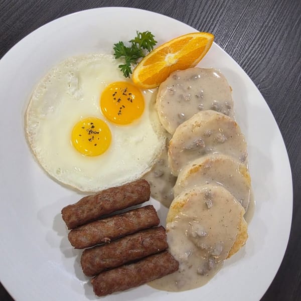 Breakfast plate with fried eggs, sausage links, biscuits with gravy, and an orange slice.