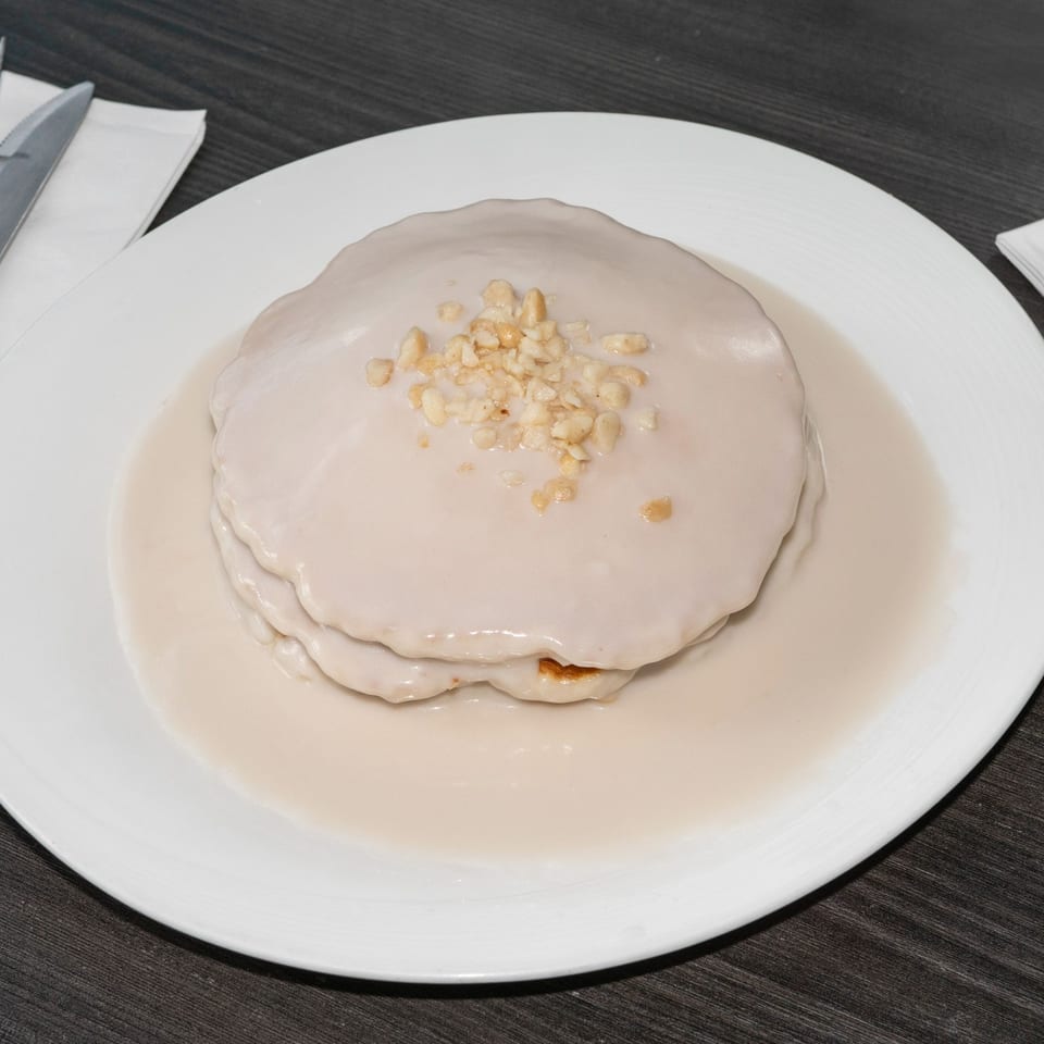 Stack of pancakes covered in white sauce and topped with chopped nuts on a white plate.