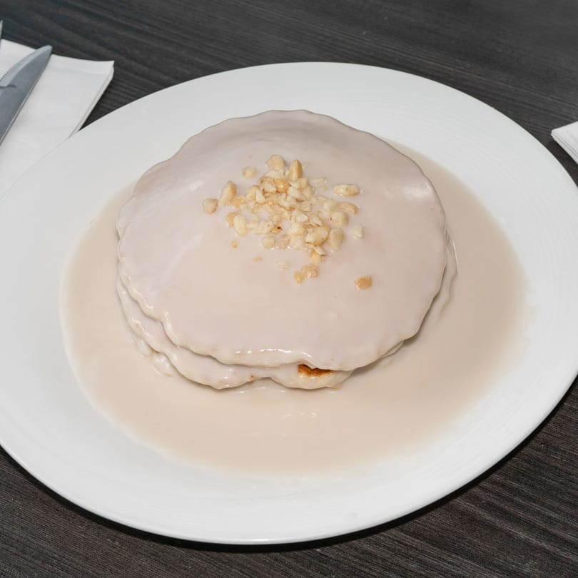 Stack of pancakes covered in white sauce and topped with chopped nuts on a white plate.