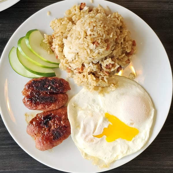 Filipino breakfast with fried rice, two fried eggs, grilled longganisa, and cucumber slices on a white plate.