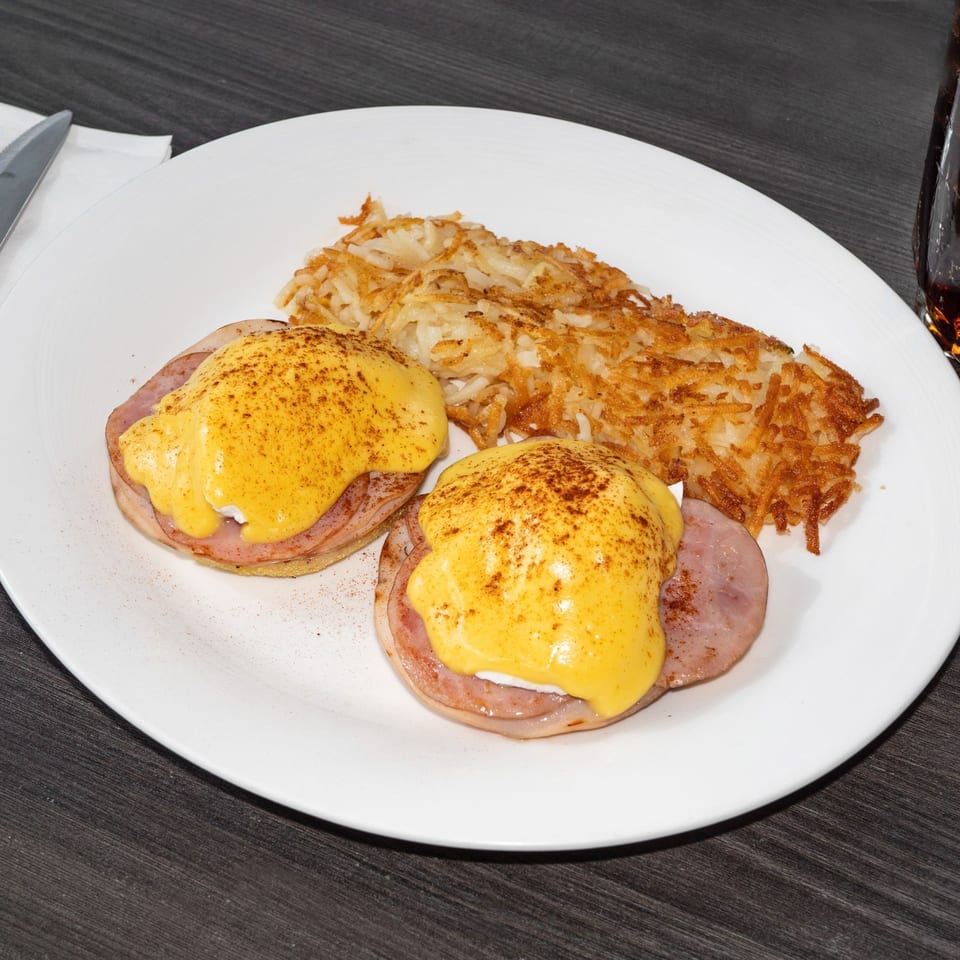 Two eggs Benedict with hollandaise sauce and hash browns on a white plate.