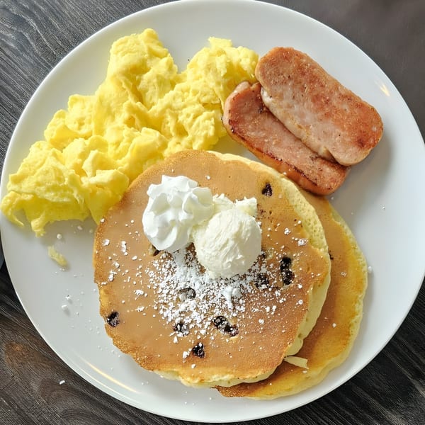 A breakfast plate with scrambled eggs, grilled spam, and blueberry pancakes topped with whipped cream.